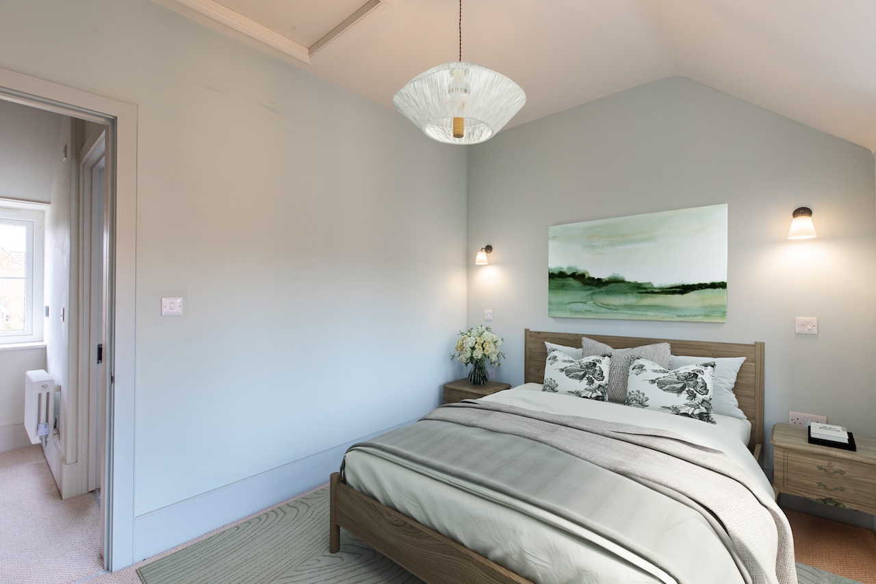 Master Bedroom Interior - Serene bedroom with soft sage green accent wall, wooden bed frame with white and patterned bedding, matching wooden nightstands, contemporary glass pendant light fixture, abstract landscape artwork in green tones, wall-mounted reading lights, white flowers in vase, en-suite bathroom visible through doorway, and neutral carpet flooring