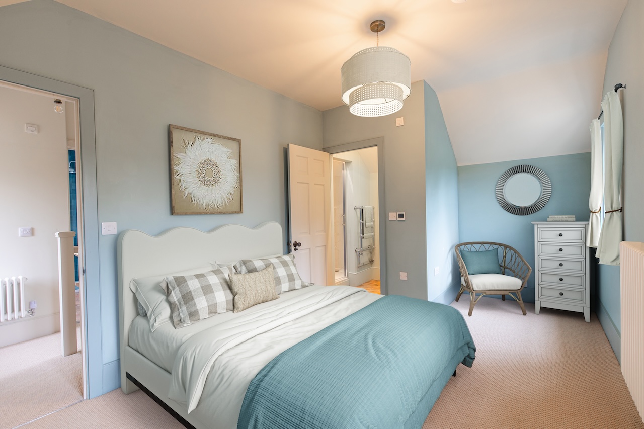 Bedroom Interior - Beautiful bedroom featuring soft teal accent wall behind white upholstered headboard, teal throw blanket, grey and white striped bedding, contemporary white pendant light with perforated design, framed sunburst artwork, white chest of drawers, wicker accent chair with teal cushion, round sunburst mirror, en-suite bathroom visible through doorway, and neutral carpet flooring