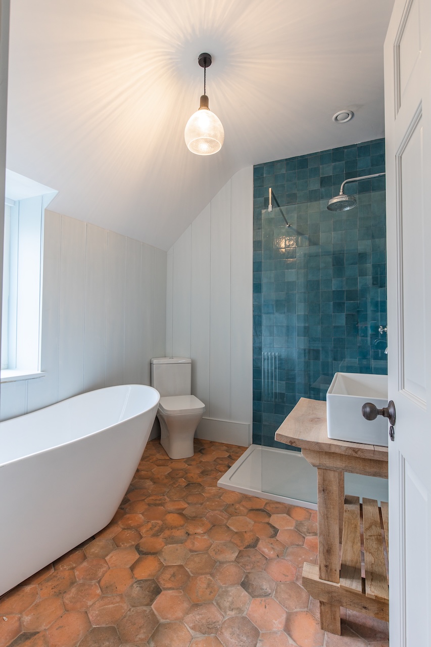 Luxury Bathroom Interior - Stunning bathroom featuring teal mosaic tile shower wall, freestanding white bathtub, modern glass pendant light, white wall paneling, terracotta hexagonal floor tiles, wooden vanity with white vessel sink, modern toilet, glass shower enclosure with rainfall showerhead, and natural light from window