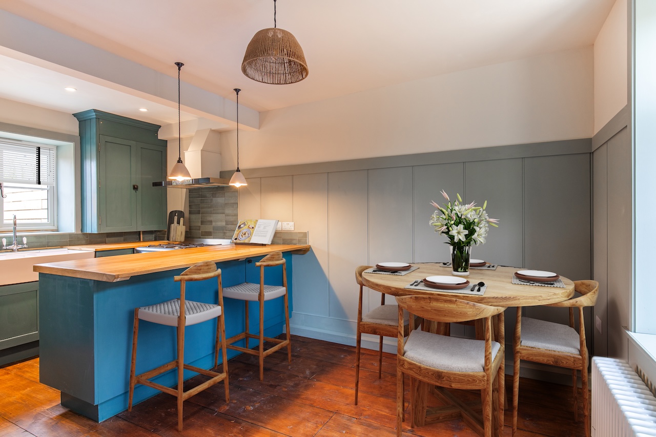 Kitchen Dining Area - Open-plan kitchen with breakfast bar seating featuring wooden bar stools with grey upholstery, round wooden dining table with matching chairs, natural wicker pendant light, exposed ceiling beams, teal kitchen island, grey wall paneling, white lilies in vase, and beautiful hardwood flooring