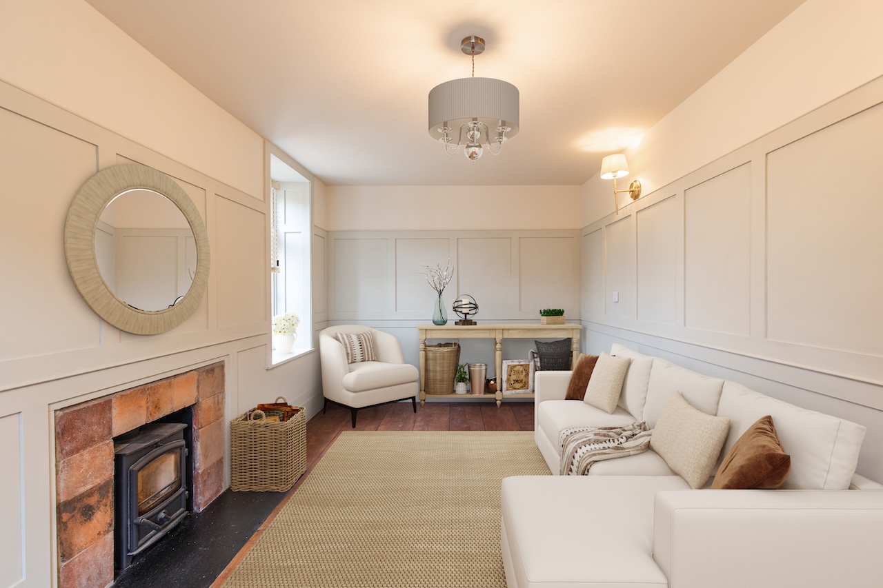 Living Room Interior - Elegant white paneled walls, exposed brick fireplace with wood-burning stove, cream L-shaped sectional sofa with brown leather and patterned cushions, round woven mirror, grey drum chandelier with crystal accents, wooden console table with decorative items, wicker storage basket, natural jute rug, and beautiful hardwood flooring