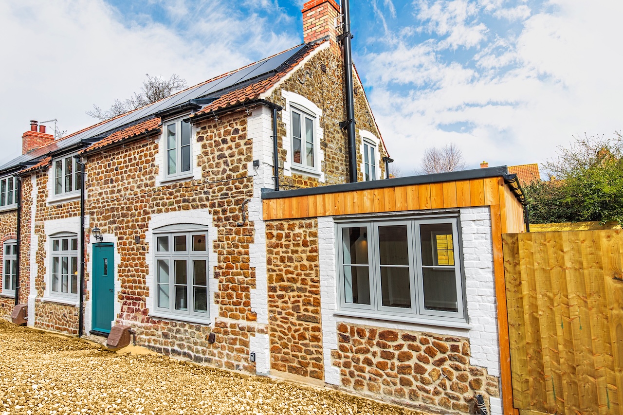 Exterior Property View - Stunning exterior showcasing traditional Norfolk flint stone construction with white mortar pointing, modern solar panels on red clay tile roof, contemporary white heritage-style windows, distinctive teal front door, modern timber-clad extension with flat roof, gravel driveway, wooden boundary fencing, and blue sky with white clouds