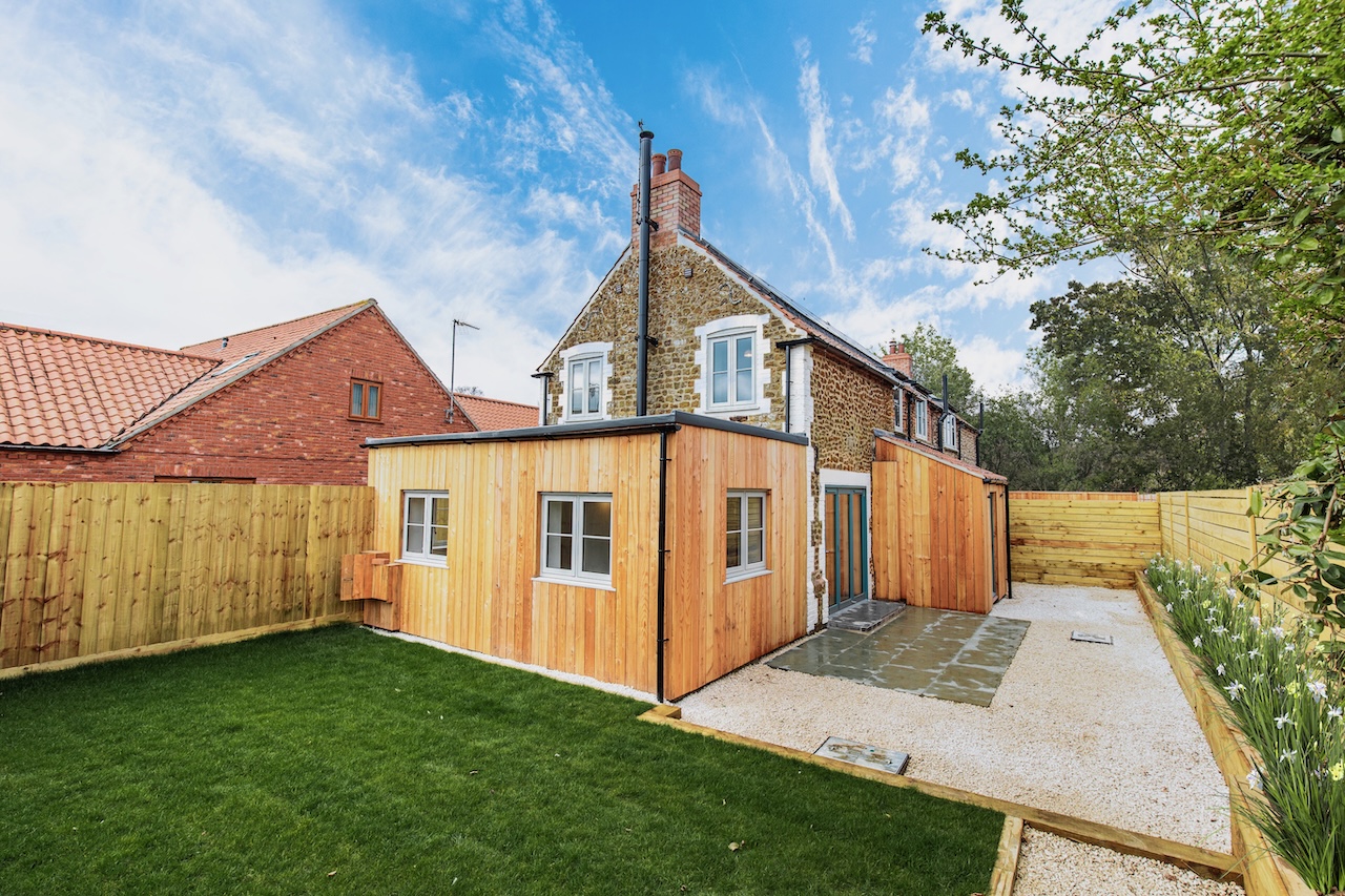 Rear Garden Exterior - Stunning rear garden image showing modern timber-clad extension with flat roof, traditional Norfolk flint stone upper level with white heritage windows, landscaped garden with fresh lawn, concrete patio area, wooden boundary fencing, mature trees, neighboring properties with red clay tile roofs, and blue sky with white clouds