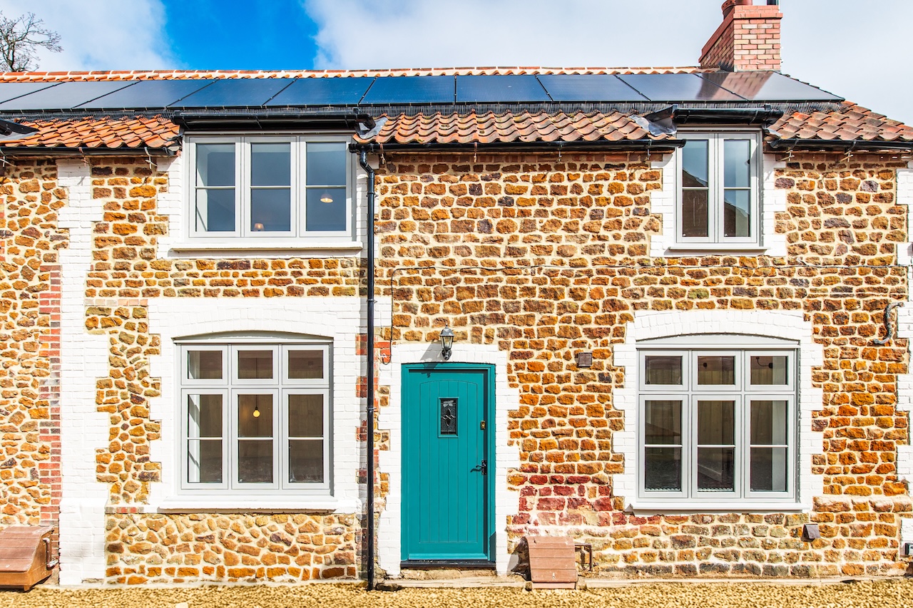 Front Elevation Exterior - Beautiful front elevation image showing traditional Norfolk flint stone construction with white mortar pointing, extensive solar panel array on red clay tile roof, contemporary white heritage-style windows with Georgian glazing bars, distinctive teal front door perfectly matching brand colors, white painted stone quoins and window surrounds, traditional lantern-style porch light, gravel driveway, and clear blue sky