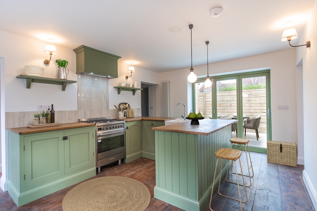 Sage Green Kitchen Interior - Beautiful traditional shaker-style kitchen featuring soft sage green cabinets with wooden countertops, white subway tile backsplash, stainless steel range cooker with extractor hood, kitchen island with breakfast bar seating, wooden bar stools with metal frames, pendant lighting, wall-mounted shelving, terracotta floor tiles, bi-fold doors to garden, and natural jute rug demonstrating exceptional craftsmanship with perfect blend of traditional and contemporary design elements