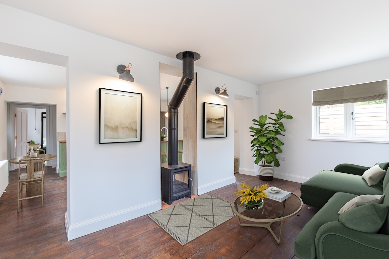 Living Room Interior - Elegant living room showing modern wood-burning stove with black flue pipe, sage green sectional sofa with cream cushions, round wooden coffee table, geometric patterned rug, abstract landscape artwork in matching frames, industrial-style wall sconces with copper accents, large fiddle leaf fig plant, Roman blind at window, open-plan connection to sage green kitchen, and beautiful dark hardwood flooring throughout