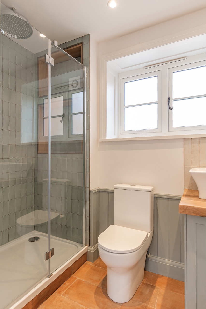 Sophisticated Bathroom Interior - Elegant bathroom featuring sage green subway tiles in shower enclosure, glass shower door with chrome fixtures, rainfall showerhead, modern white toilet, sage green tongue-and-groove wall paneling, wooden vanity countertop with white vessel sink, terracotta floor tiles, white casement window with Georgian glazing bars, and recessed ceiling lighting perfectly matching the property's sage green color scheme with high-quality traditional materials