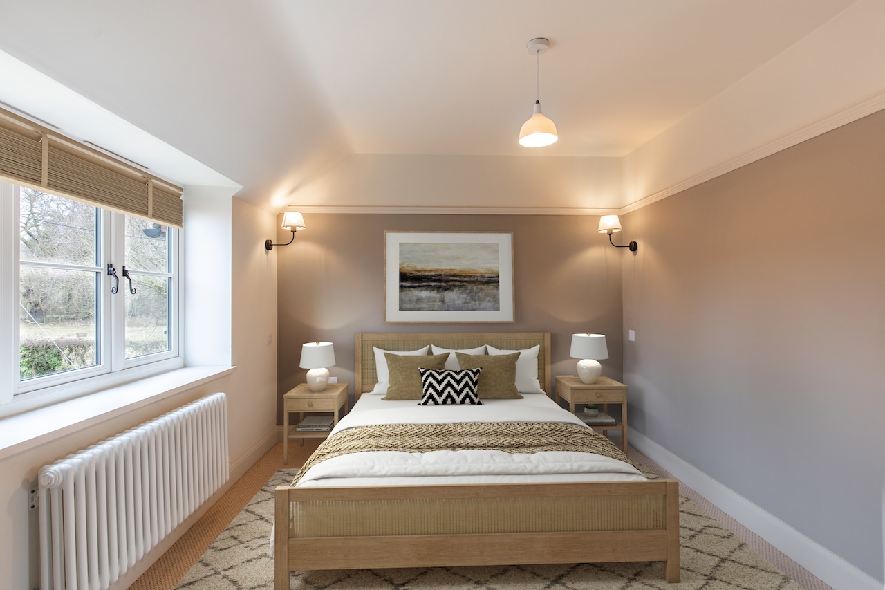 Bedroom Interior - Sophisticated bedroom image showing warm taupe accent wall behind wooden bed frame with cane headboard detail, white bedding with olive green cushions and chevron patterned pillow, woven throw blanket, matching wooden nightstands with white ceramic table lamps, wall-mounted reading lights with white shades, abstract landscape artwork in white frame, pendant ceiling light, Roman blind at large window, white column radiator, and neutral carpet with geometric patterned rug demonstrating elegant interior design with natural materials and warm neutral tones